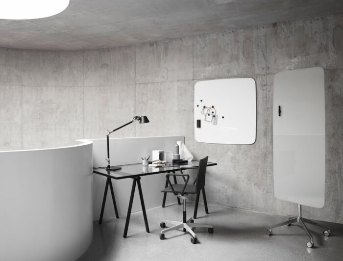 Flow Mobile Writing Board with soft curved edges in white finish with polished aluminium base on castors shown in a work space with a desk and glass wall board